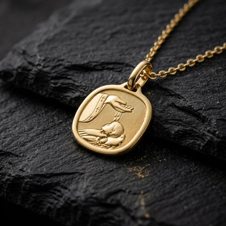 Yellow gold rounded-square baptism medal pendant with a polished rim and a raised priest's hand pouring baptism water over an infant, 14.6 mm, on a yellow gold chain resting on dark slate stone