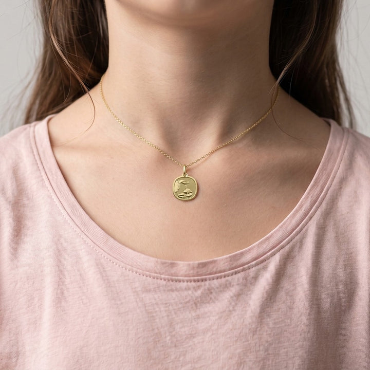 Yellow gold rounded-square baptism medal pendant with a polished rim and a raised priest's hand pouring baptism water over an infant, 14.6 mm, worn by a girl on a fine yellow gold chain