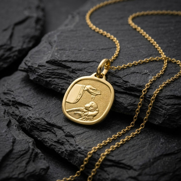 Yellow gold rounded square baptism medal pendant with a wide polished bevelled rim and a raised hand pouring water over a sleeping infant beside a priest's stole with crosses, 16 mm wide, on a yellow gold chain resting on dark slate stone