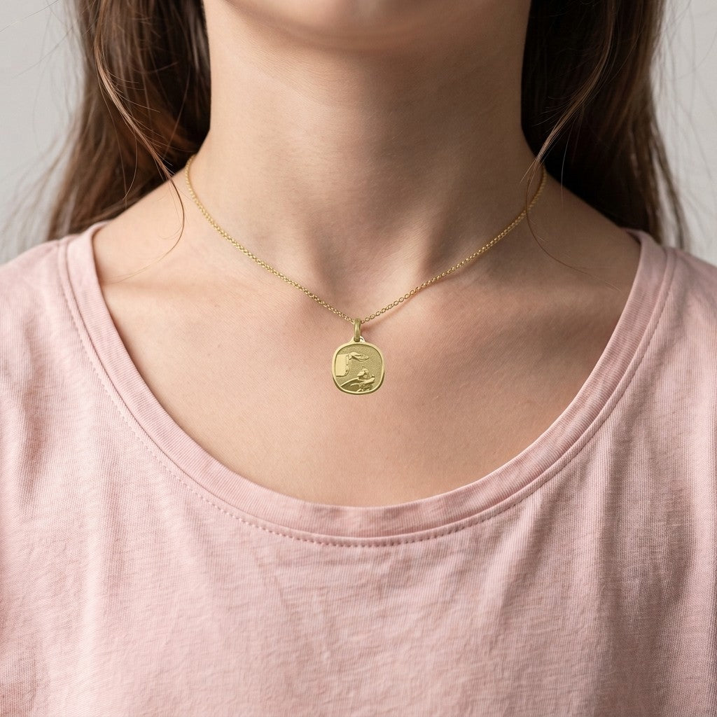 Yellow gold rounded square baptism medal pendant with a wide polished bevelled rim and a raised hand pouring water over a sleeping infant beside a priest's stole with crosses, 16 mm wide, worn by a girl on a fine yellow gold chain