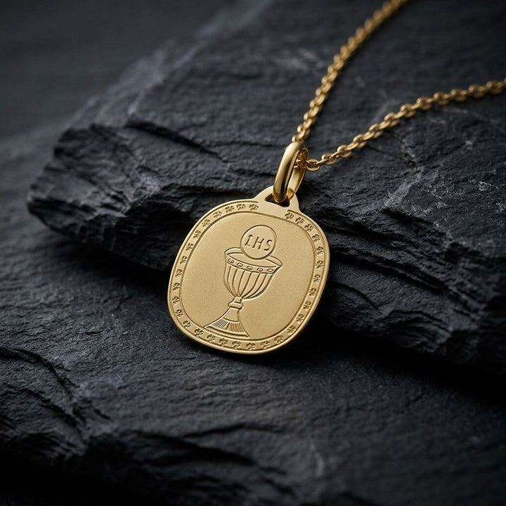 Yellow gold rounded square communion medal pendant with an engraved chalice and host inside a clover-patterned border, 16.2 mm wide, on a yellow gold chain resting on dark slate stone