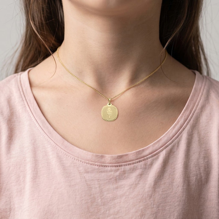 Yellow gold rounded square communion medal pendant with an engraved chalice and host inside a clover-patterned border, 16.2 mm wide, worn by a girl on a fine yellow gold chain