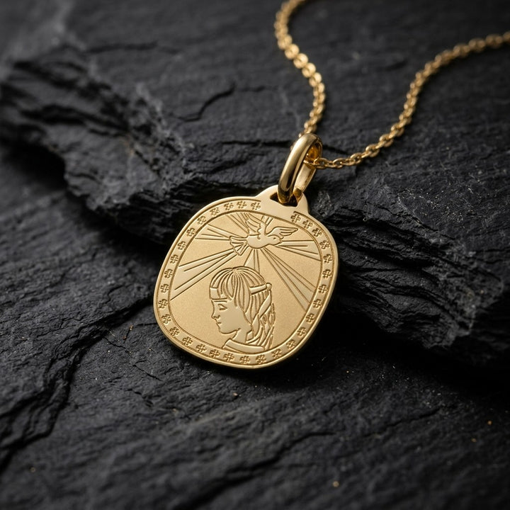 Yellow gold rounded square confirmation medal pendant with an engraved dove sending rays of light onto a praying girl inside a clover-patterned border, 16.2 mm wide, on a yellow gold chain resting on dark slate stone