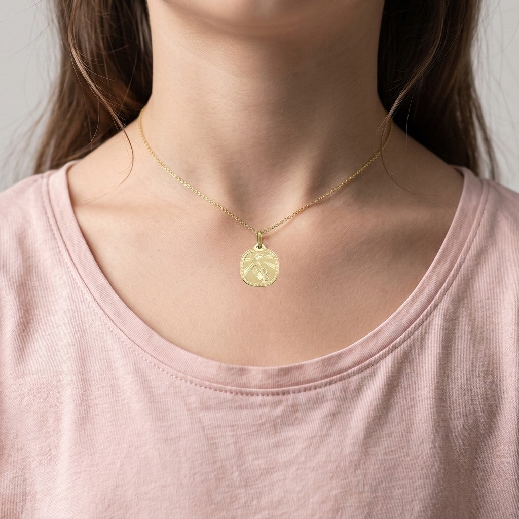 Yellow gold rounded square confirmation medal pendant with an engraved dove sending rays of light onto a praying girl inside a clover-patterned border, 16.2 mm wide, worn by a girl on a fine yellow gold chain