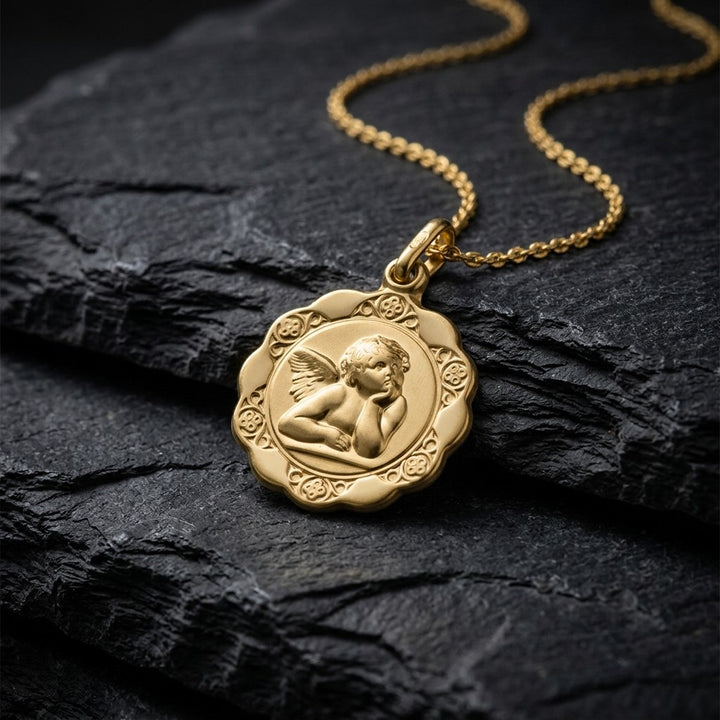 Yellow gold scalloped angel medal pendant with a raised Raphael cherub in relief and a rosette border, 16.8 mm, on a yellow gold chain resting on dark slate stone