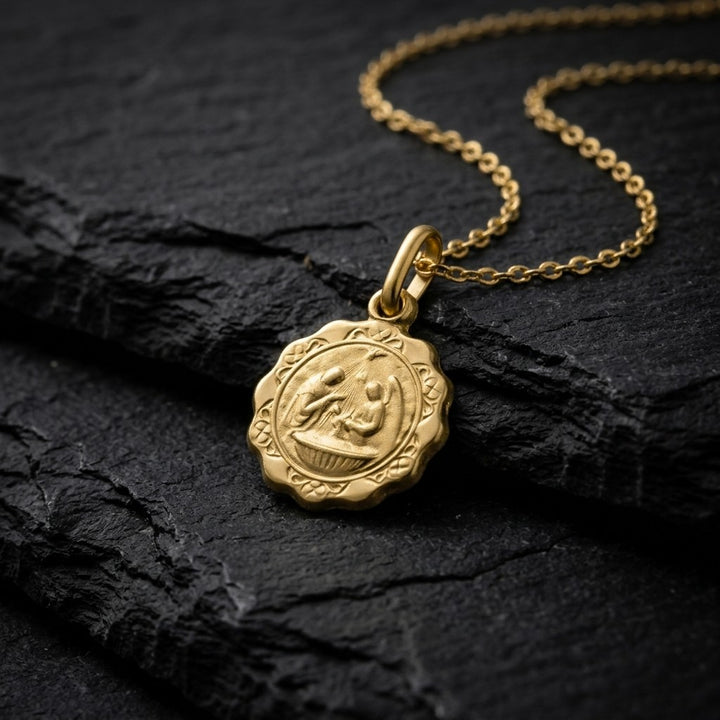 Yellow gold scalloped baptism medal pendant with a flower-shaped lobed edge, a scroll border and a raised priest, angel and infant at the font scene, 12.4 mm, on a yellow gold chain resting on dark slate stone