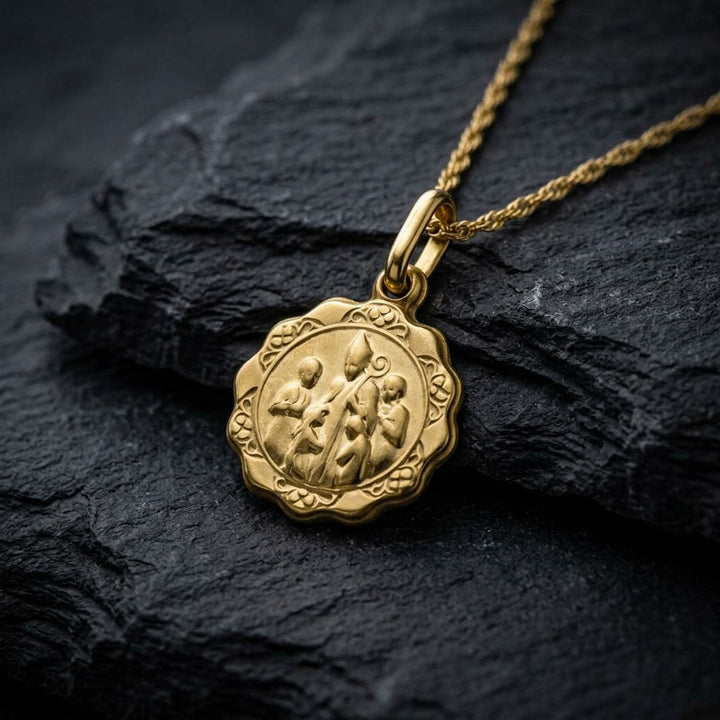 Yellow gold scalloped confirmation medal pendant with a bishop in a tall mitre laying his hand on a kneeling child, standing children at each side and rosette ornaments around the border, 12.4 mm wide, on a yellow gold chain resting on dark slate stone