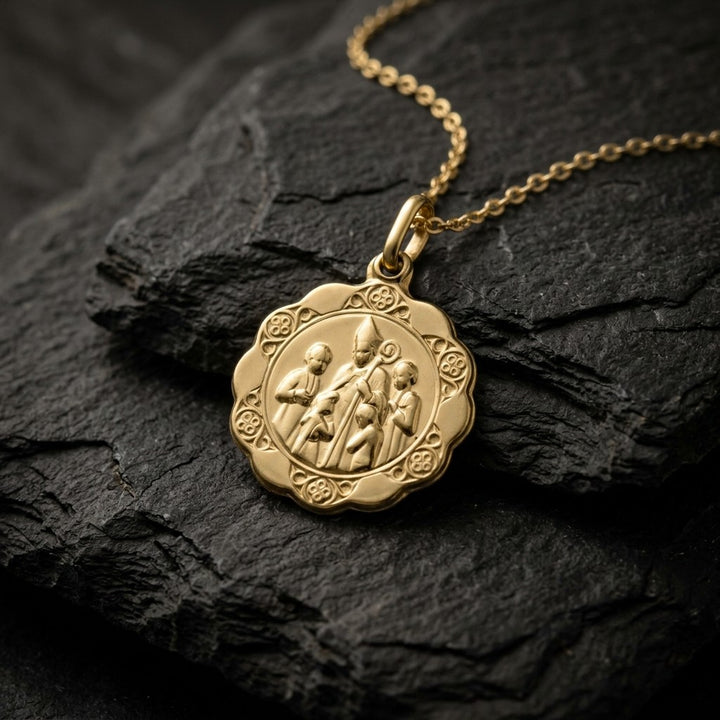 Yellow gold scalloped confirmation medal pendant with a bishop in a tall mitre laying his hand on a kneeling child, standing children at each side and rosette ornaments around the border, 16.8 mm wide, on a yellow gold chain resting on dark slate stone