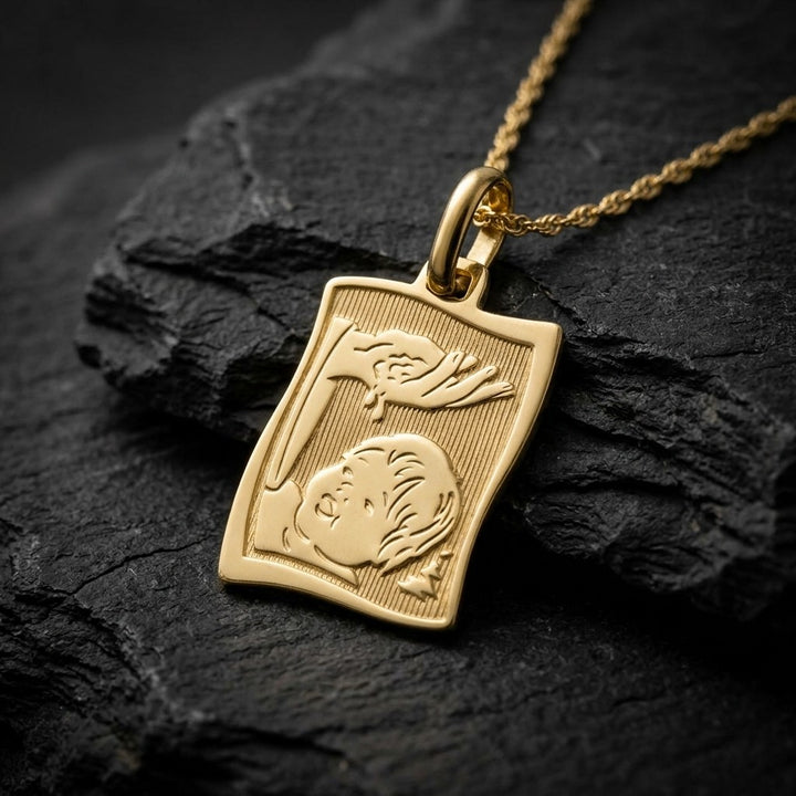 Yellow gold wavy rectangle scroll-style baptism medal pendant with a ribbed background and a raised hand pouring water over an infant's face, 13 mm wide, on a yellow gold chain resting on dark slate stone